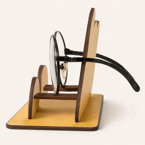 May include: A light-colored wooden eyeglass stand with a flat base and a vertical back panel featuring rounded edges. A pair of black-framed glasses rests horizontally on the small wooden support shelf extending from the front of the stand.