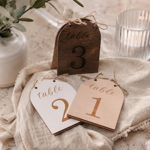 May include: Three arched wooden table signs featuring the text table and numbers 1, 2, and 3. The signs are made of wood in dark brown, white, and light natural finishes, each topped with a small decorative jute twine bow tied through two holes.