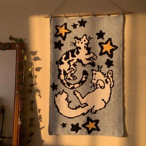 Peut inclure: Une tenture murale rectangulaire en crochet bleu clair représentant deux chats blancs avec des taches noires, entourés d'étoiles noires et jaunes. Le textile est suspendu à une tige en bois avec une ficelle, affichant un style pixelisé.
