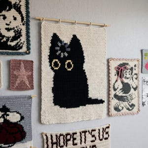 Peut inclure: Une tenture murale rectangulaire en crochet couleur crème représentant la silhouette d'un chat noir avec de grands yeux jaunes et une petite fleur grise sur la tête, suspendue à une fine tige en bois avec des boucles sur le bord supérieur.