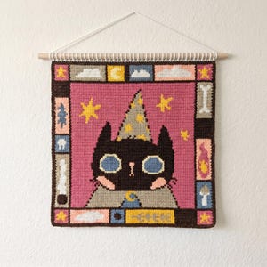 Op de afbeelding: Een vierkante gehaakte wandhanger met een zwarte kat die een puntmuts met gele sterren en een grijze cape draagt tegen een roze achtergrond. De rand bevat kleine rechthoekige panelen met iconen van sterren, wolken, een maan, botten en paddenstoelen.