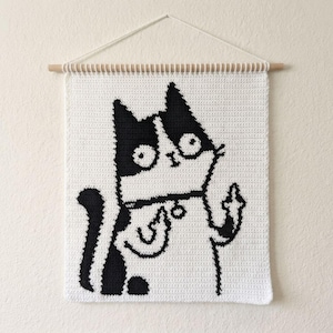 Puede incluir: Un tapiz de ganchillo rectangular con un gráfico en blanco y negro de un gato con ojos grandes y un collar. El gato aparece con una pata levantada, mostrando un gesto con el dedo corazón. La pieza está suspendida de un listón de madera claro.