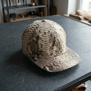 May include: A baseball cap featuring a textured, scale-like pattern in shades of beige, grey, and dark brown. The cap has a rounded crown with small metal eyelets for ventilation and a flat brim, showcasing a detailed, reptilian-inspired surface.