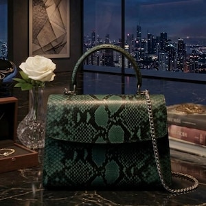 May include: A structured handbag featuring a dark green and black snakeskin pattern. The bag has a single top handle, a fold-over flap closure, and a detachable silver-toned metal chain shoulder strap. The exterior texture mimics reptile scales.