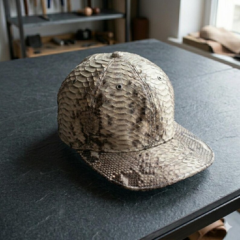 Pu&ograve; includere: Un cappellino da baseball caratterizzato da un motivo testurizzato simile a scaglie nei toni del beige, grigio e marrone scuro. Il cappellino presenta una corona arrotondata con piccoli occhielli metallici per l'aerazione e una visiera piatta.