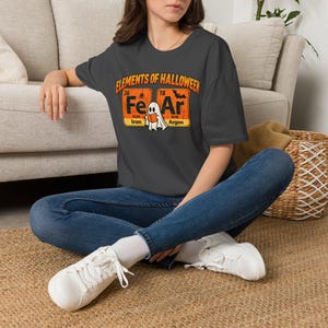 Könnte beinhalten: Ein dunkelgraues T-Shirt mit einem Grafikdruck, der Elements of Halloween liest. Das Design zeigt zwei Periodensystem-Quadrate für Eisen und Argon, mit einem weißen Geist, der ein orangefarbenes Buch dazwischen hält.