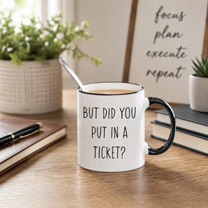 May include: A white ceramic mug with a black rim and handle, featuring the black text "BUT DID YOU PUT IN A TICKET?" printed on the front. The mug contains a light brown liquid and a silver spoon is resting inside the vessel on a wooden surface.