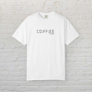 May include: A white short-sleeve t-shirt featuring a graphic on the chest with the text CO:FF:EE TIME written in a digital clock font style. The shirt has a crew neckline and a standard fit, displaying the text clearly in dark gray ink.