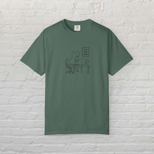 May include: A sage green short-sleeve t-shirt featuring a black line art graphic of a pixelated dinosaur sitting at a desk with a laptop and a mug. Above the desk, a framed sign displays the text JUST KEEP RUNNING in a simple block font.