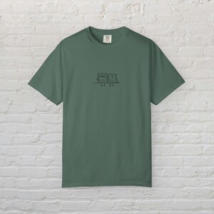 Pode incluir: Uma t-shirt de manga curta verde-sálvia com um pequeno gráfico de linhas pretas centrado no peito. O gráfico mostra duas teclas de teclado de computador com as etiquetas Ctrl e Z, cada uma com braços e pernas simples sentadas numa linha.