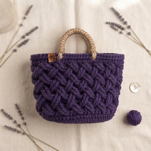 May include: A deep purple crocheted basket with a thick, braided texture and a single tan jute handle. A small rectangular brown leather tag with the text Mela is attached to the upper left side of the basket, which features a woven pattern.