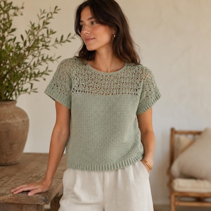 May include: A sage green short-sleeved knit top featuring a round neckline and a decorative openwork lace pattern across the upper chest and shoulders. The garment has a solid, dense stitch texture on the lower body and ribbed hem detailing.