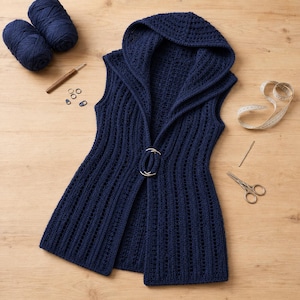 May include: A dark navy blue sleeveless hooded vest with a vertical ribbed crochet pattern, featuring a large silver-toned circular metal clasp at the centre. Two balls of matching navy yarn, a crochet hook, stitch markers, and scissors are included.