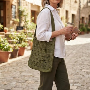 May include: An olive green crochet tote bag with a rectangular shape and an open-weave pattern. The bag features a single wide shoulder strap and is constructed from yarn, showcasing a textured, handmade appearance with a consistent stitch design.
