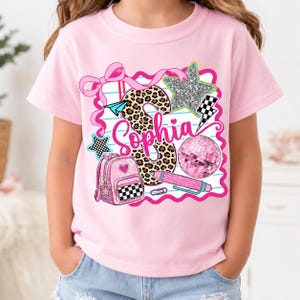 Pode incluir: Uma t-shirt rosa claro com um design gráfico que apresenta o nome Sophia em letra cursiva. A estampa inclui uma letra S com padrão de leopardo, uma mochila rosa, uma bola de discoteca, um lápis, um avião de papel, estrelas e um laço rosa.