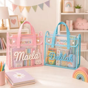 May include: Two rectangular clear plastic tote bags with fabric handles and trim. The left bag has pink trim and the name Maela in white script. The right bag has blue trim and the name Delilah in blue script. Both bags contain school supplies.