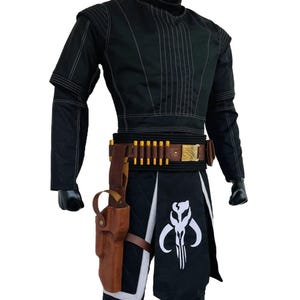 May include: A black long-sleeved tunic and pants set featuring white vertical stitching, a brown leather utility belt with gold-colored ammunition cartridges, a matching brown leather holster, and a black loincloth with a white skull emblem.