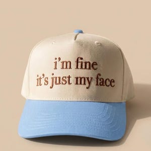 May include: A beige baseball cap with a light blue brim. The front of the cap features brown embroidered text that reads, "i'm fine it's just my face." The cap has a structured crown with a small blue button at the top and stitched detailing.