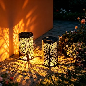 Peut inclure: Deux lanternes cylindriques en métal noir avec des découpes complexes en forme de branches d'arbre. Chaque lanterne possède un panneau solaire sur le dessus et une poignée en fil de fer, diffusant une lumière chaude et projetant des ombres.
