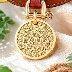 May include: A circular gold-toned metal pet identification tag featuring an engraved pattern of overlapping fan-like shapes and the name Coco centered in bold lettering. The tag is attached to a brown leather collar by a gold-toned metal shackle.