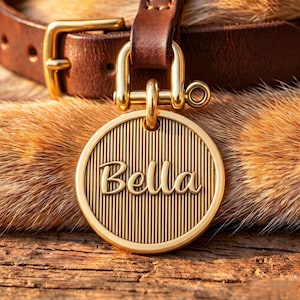 Op de afbeelding: Een ronde, goudkleurige metalen huisdierpenning met een verticale ribbeltextuur en de naam Bella gegraveerd in een cursief lettertype. De penning is met een goudkleurige sluiting bevestigd aan een donkerbruine leren halsband met metalen gesp.