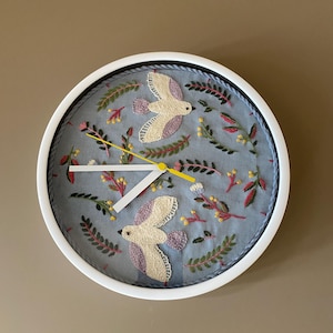 Peut inclure: Une horloge murale ronde avec un cadre blanc et un cadran en tissu bleu clair orné d'oiseaux brodés à la main en crème et violet, entourés de petites tiges florales vertes, roses et jaunes. Elle possède deux aiguilles blanches et une trotteuse jaune.