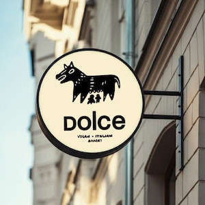 May include: A circular, illuminated sign mounted on a building wall. The sign features a cream-colored background with a black graphic of a stylized animal and the text "dolce" followed by "VEGAN + ITALIAN BAKERY" in black lettering.