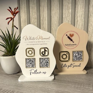 May include: Two irregular-shaped acrylic signs with stands. The white sign features the text White Moment, Instagram and TikTok icons, and two QR codes with the text Follow us. The beige sign features a NabsSweets logo, an Instagram icon, a QR code, and the text Let's get Social.