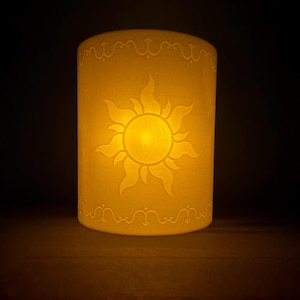 Puede incluir: Una lámpara cilíndrica de color blanco translúcido con un emblema de sol amarillo brillante en el centro. El diseño incluye bordes de filigrana decorativos en la parte superior e inferior, emitiendo una luz cálida y suave sobre un fondo oscuro.