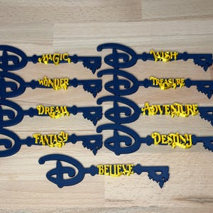 May include: A collection of nine dark blue decorative keys featuring the iconic Disney logo as the bow. Each key displays a unique yellow word: Magic, Wish, Wonder, Treasure, Dream, Adventure, Fantasy, Destiny, and Believe, arranged in a grid.
