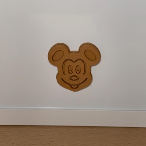 Puede incluir: Un emblema de plástico moldeado, plano y de color marrón claro, que presenta la cabeza y el rostro icónicos de un ratón de dibujos animados. El diseño incluye orejas redondeadas, ojos ovalados, una nariz pequeña y una boca ancha y sonriente.