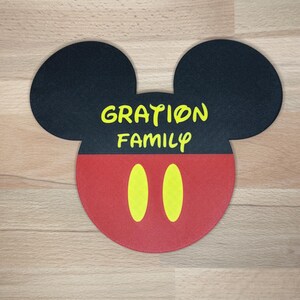 May include: A flat, circular-based coaster shaped like a mouse head with black ears and a red bottom section. The black area features the yellow text "GRATION FAMILY" in a stylised font, and the red section has two vertical yellow oval shapes.