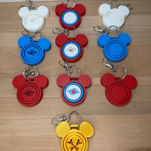 May include: A collection of ten Mickey Mouse-shaped keychains in various colours including white, red, blue, and yellow. Each keychain features a circular centre with different embossed designs, including cruise ship logos and crossed keys, attached to a metal ring.