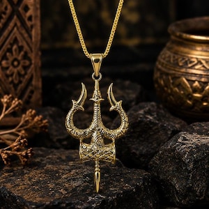 Puede incluir: Un colgante de metal dorado con forma de tridente, que presenta intrincados patrones grabados y una base cilíndrica central, suspendido de una cadena a juego. El colgante tiene tres puntas y un extremo inferior afilado, con un acabado pulido.