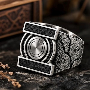 May include: A silver-toned metal ring featuring a central circular emblem with concentric rings, flanked by two horizontal rectangular bars with a black textured finish. The band has a rugged, weathered surface with deep cracks and dark detailing.
