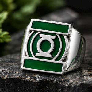 May include: A silver-toned ring featuring a prominent rectangular face with a central circular Green Lantern emblem. The design includes a vibrant green enamel inlay surrounding the silver symbol and two horizontal green bars at the top and bottom.