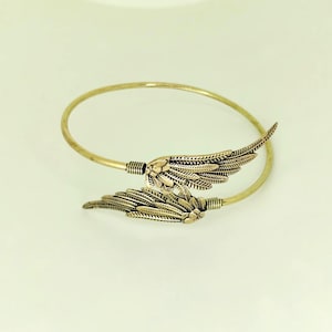 May include: A gold-toned metal bangle bracelet featuring two overlapping wing-shaped charms. The wings have detailed, etched feather textures and are connected to the thin, circular band by small, ribbed cylindrical metal components.