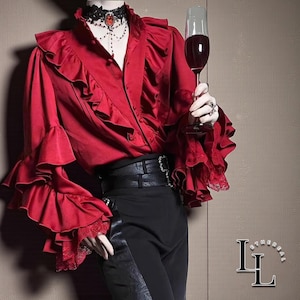 Op de afbeelding: Een rood shirt met lange mouwen en ruches aan de voorzijde en mouwen, voorzien van zwarte kanten afwerking bij de manchetten. Het shirt wordt gecombineerd met een zwarte broek en een brede zwarte riem. Een zwarte kanten choker met een rode steen.