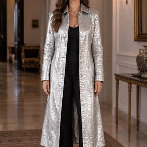 Puede incluir: Un abrigo largo de color plateado con un patrón texturizado en relieve que imita la piel de cocodrilo. El abrigo cuenta con un cuello clásico, mangas largas y cierre frontal de botones. Se lleva sobre una camiseta negra y pantalones negros.