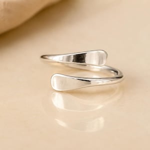 Può includere: Un anello aperto in tonalità argento lucido caratterizzato da un design liscio con estremità arrotondate e appiattite. La superficie metallica è riflettente e continua, curvandosi per formare una struttura a fascia regolabile.