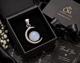 Puede incluir: Un colgante de media luna de plata con un cabochón de piedra lunar iridiscente redondo en el centro. El colgante se muestra dentro de un joyero cuadrado negro con un lazo de cinta negra en la tapa y una tarjeta que dice Thank You.