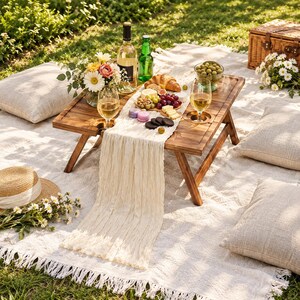 May include: A rectangular wooden folding picnic table with integrated wine glass holders, topped with a cream-colored cheesecloth runner, a bottle of white wine, a green bottle, a glass of wine, and a charcuterie board with fruit and pastries.