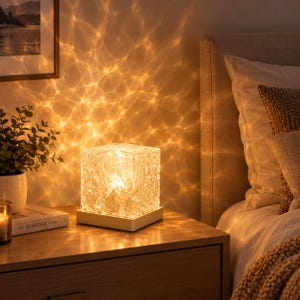 May include: A square, clear glass lamp with a textured, ice-like surface sits on a flat, light-colored base. When illuminated, the lamp casts a warm, golden, rippling light pattern onto the surrounding wall, creating a water-like effect.