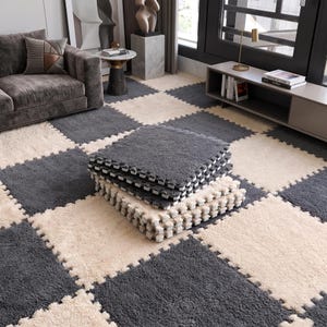 May include: A stack of square interlocking floor tiles featuring a plush, shaggy texture in alternating dark gray and cream colors. The tiles are designed with puzzle-piece edges that fit together to create a checkered pattern on the floor.