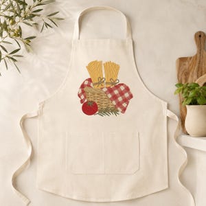 May include: A cream-colored apron featuring a central graphic print of a woven basket filled with two bundles of yellow spaghetti, a red tomato, and green herbs, all draped with a red and white checkered cloth. The apron includes a front pocket.