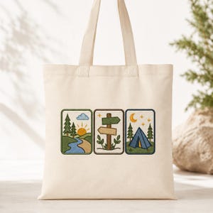 Op de afbeelding: Een crèmekleurige canvas draagtas met drie rechthoekige geborduurde patches. De ontwerpen tonen een zonsopgang boven een rivier en bomen, een houten wegwijzer met twee pijlen en een blauwe tent onder een halve maan en sterren.