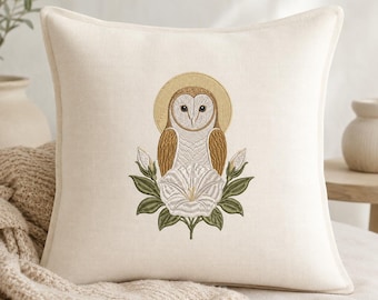 May include: A square off-white throw cushion featuring a central embroidered design of a barn owl with a golden halo, perched atop a large white morning glory flower with green leaves and two closed flower buds, centred on the fabric.