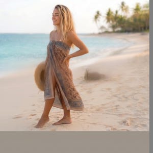 Puede incluir: Un pareo sin tirantes con un estampado floral en tonos marr&oacute;n y azul con un borde decorativo. La tela ligera est&aacute; envuelta alrededor del torso y las piernas, creando una silueta fluida. Se sostiene un sombrero de paja de ala ancha a un lado.