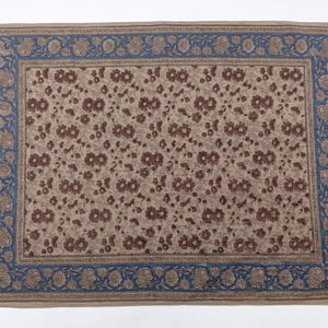 Puede incluir: Un textil rectangular que presenta un campo central de patrones florales marrones sobre un fondo beige claro, encerrado por un borde ancho de motivos florales azules y marrones. La tela muestra un dise&ntilde;o de estampado en bloque.