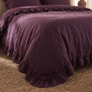 Puede incluir: Una funda nórdica de color ciruela intenso con un borde de volantes a lo largo de todo el perímetro. La tela tiene un tejido texturizado visible y un acabado mate suave. El juego de cama incluye fundas de almohada a juego con volantes.
